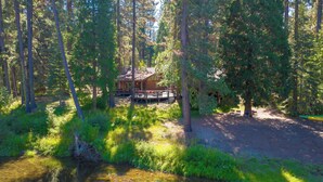 Une cabane entourée d’une forêt dense, un ruisseau aux eaux claires au premier plan et une terrasse avec des chaises.