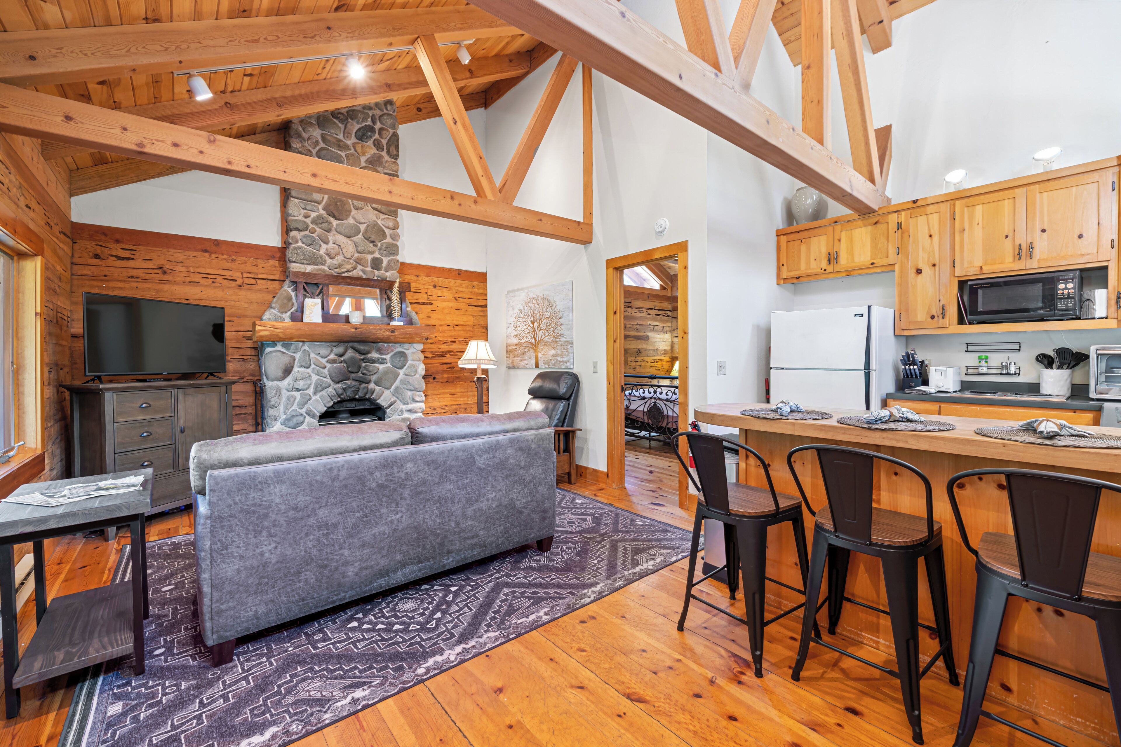 A spacious living area with a stone fireplace, wooden beams, a grey sofa, and a kitchen with wooden cabinets and a bar area.