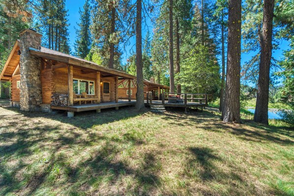A rustic cabin with a stone chimney, wooden deck, and a view of a lake surrounded by tall pine trees. - 2 Riverfront, Dog-friendly Cabins on 200 Acres (Blairsden-Graeagle)