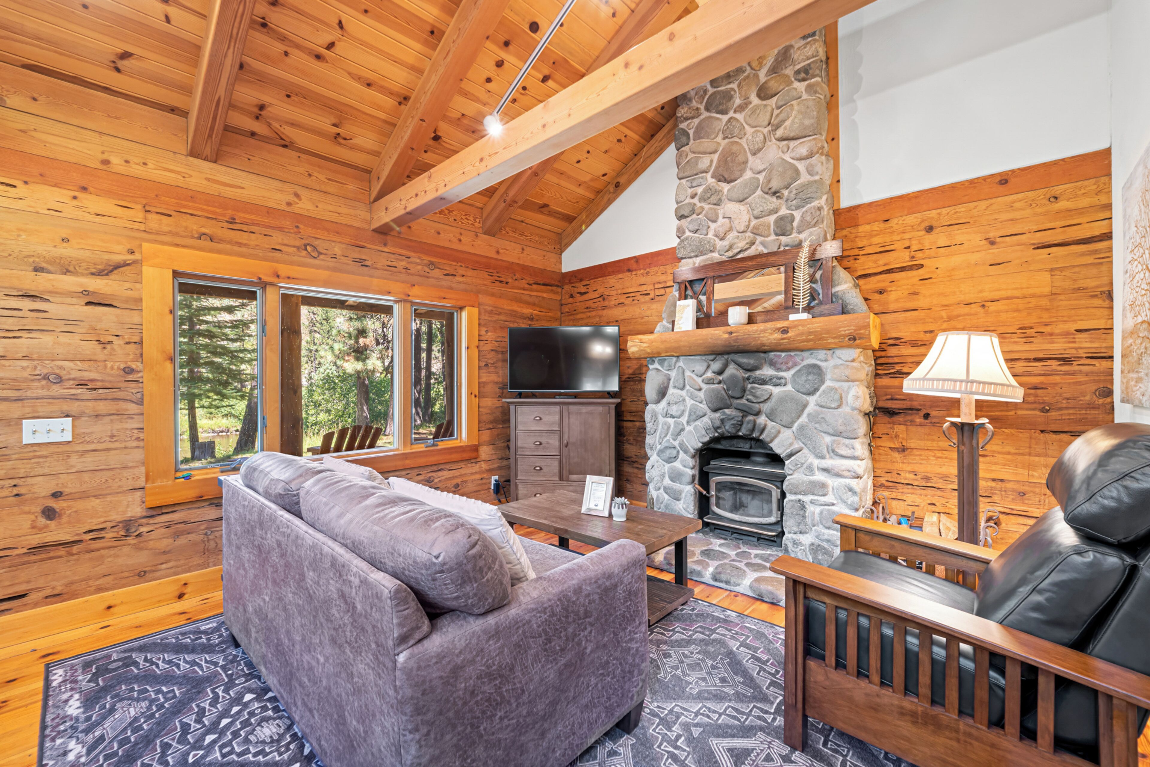A cozy cabin interior with a stone fireplace, wooden ceiling beams, a sofa, and a television.
