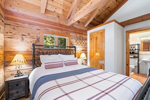 A wooden cabin bedroom with a bed, two bedside lamps, a window, and a door leading to a bathroom. - 2 Riverfront, Dog-friendly Cabins on 200 Acres (Blairsden-Graeagle)