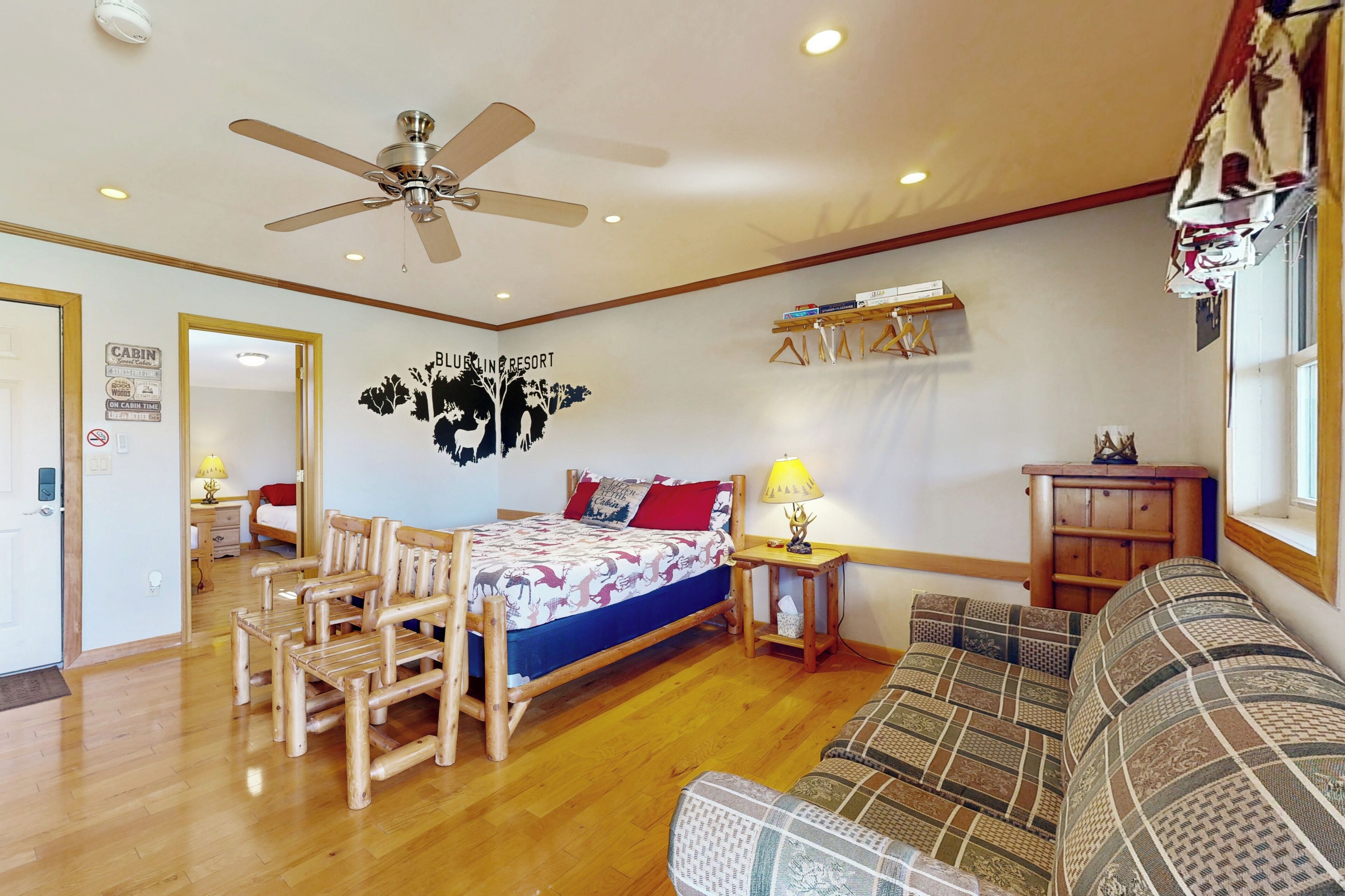 A bedroom with a bed, wooden chairs, a sofa, a ceiling fan, a nightstand, a lamp, and a wall decoration.