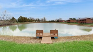 A serene outdoor setting with a wooden dock leading to a calm pond, surrounded by a grassy area and a few buildings in the background. - Blue Line Resort - All 3 Cabins! (Murphysboro)