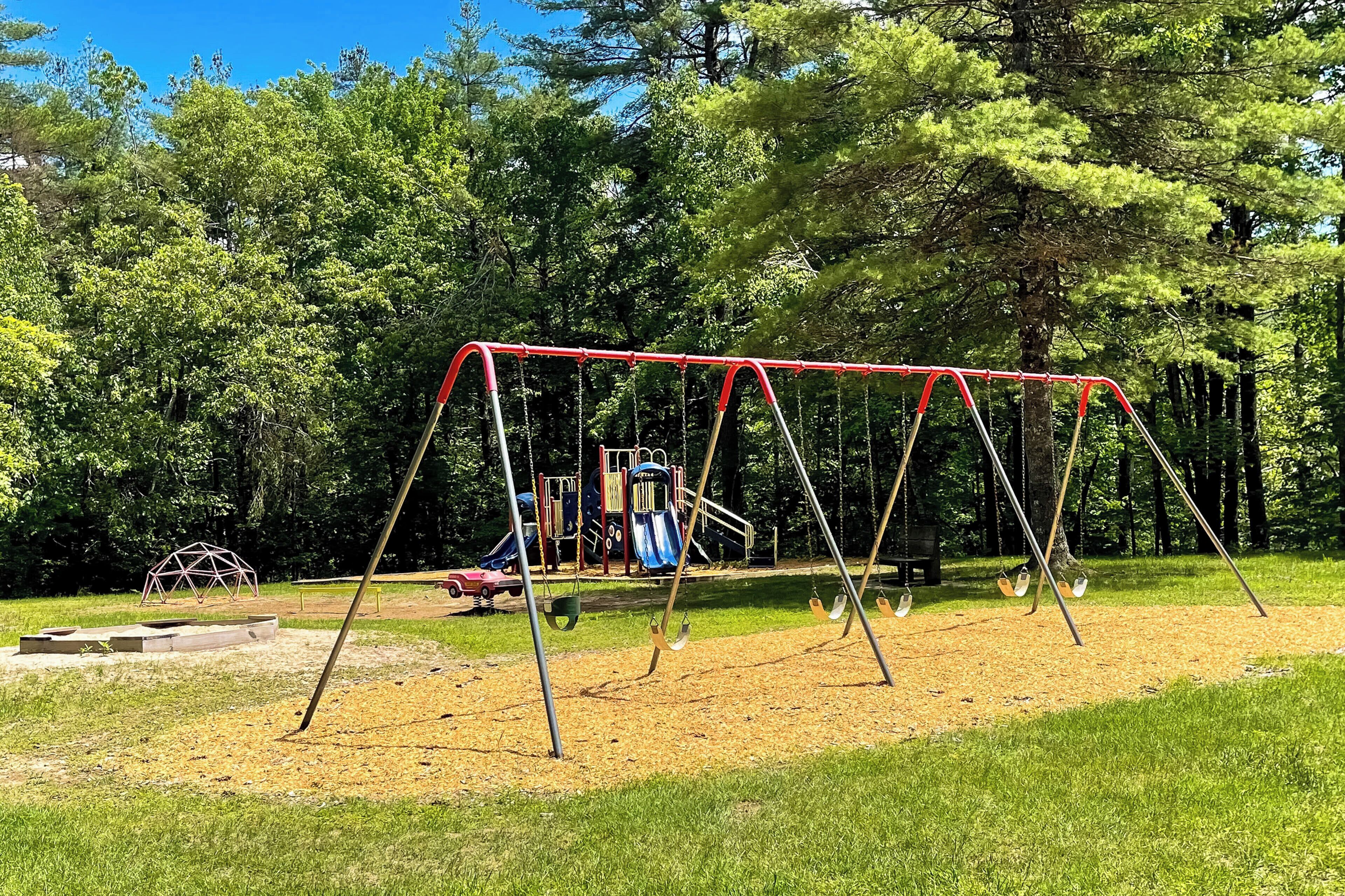 Un parque infantil con columpios y un tobogán, rodeado de árboles y un cielo despejado.