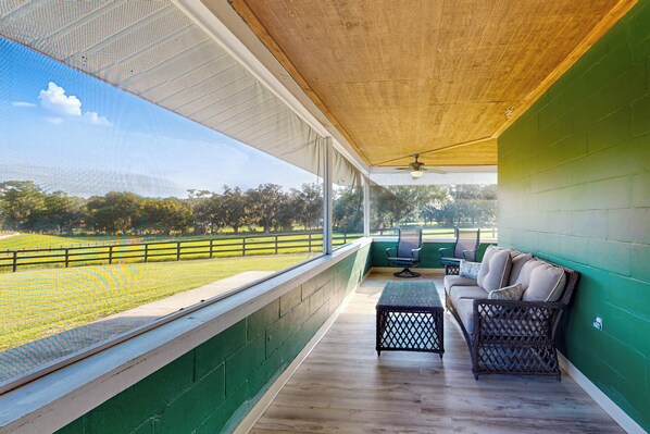 A covered outdoor seating area with a view of a sports field, a wicker sofa, and a small table. - Charming Two Springs Farm Guesthouse! (Micanopy)