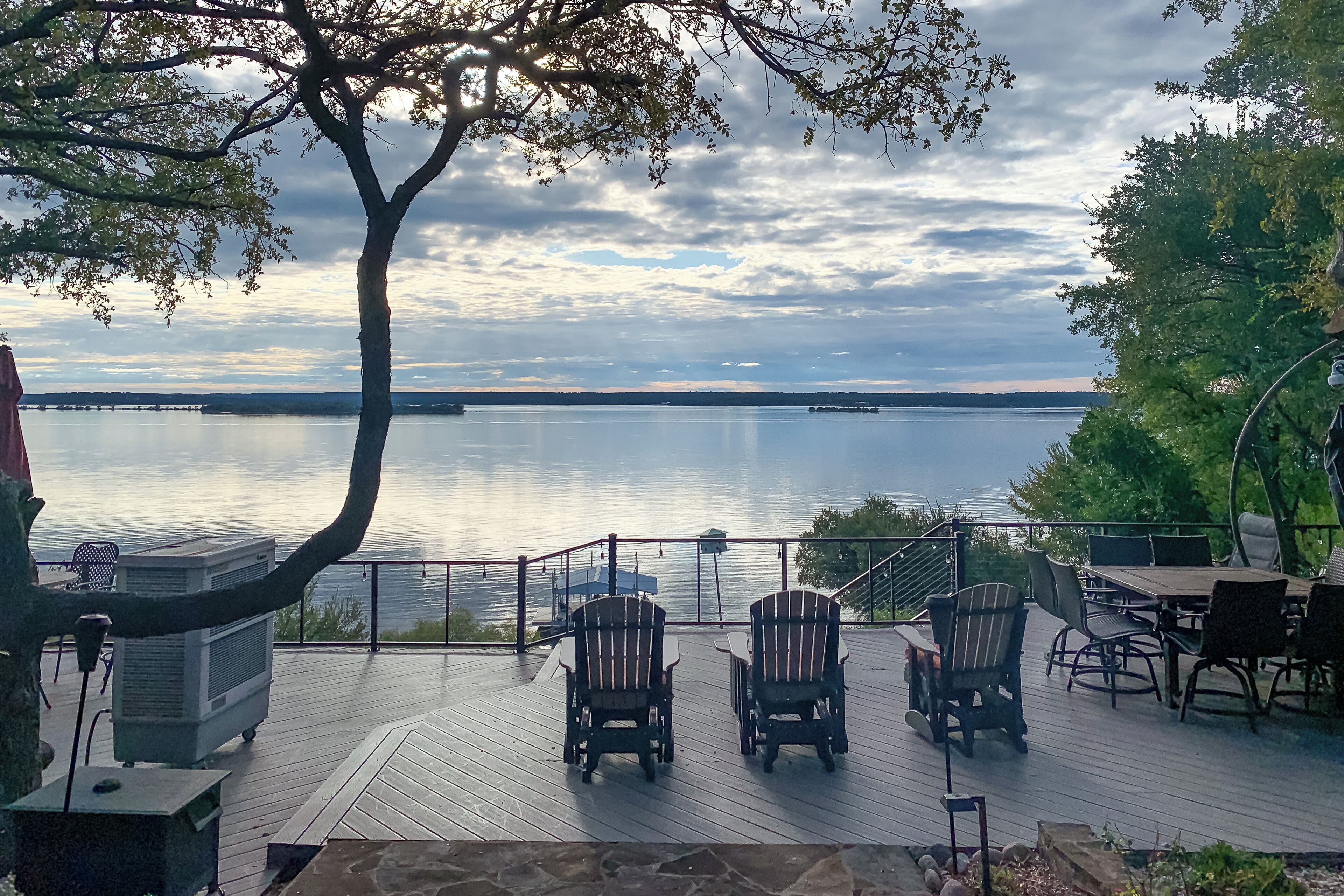 A deck with Adirondack chairs overlooking a lake, a distant bridge, and a cloudy sky.