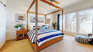A four-poster bed with a striped bedspread, a wooden nightstand with a lamp, and a view of the outdoors through large windows. - Lakefront Estate: Stunning Views, Deck, Bar & Dock (Bridgeport)
