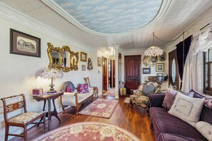 A room with a wooden floor, a purple sofa, a wooden chair, a table with a lamp, and a mirror with a gold frame.