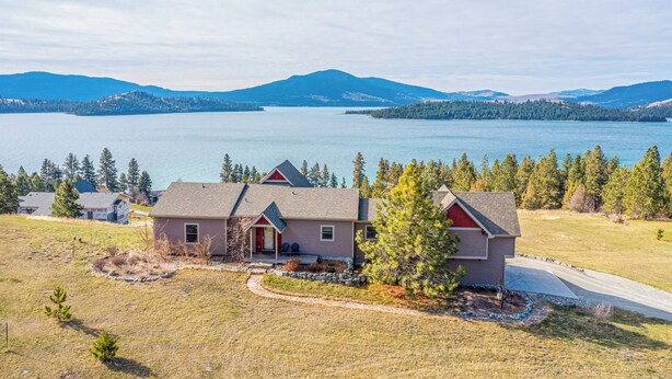 A single-story house with a red front door, a grey roof, and a small porch. It is surrounded by a well-maintained lawn and a few trees. In the background, there is a large body of water and a mountain range. - Breathtaking Mountain & Lake Views! (Dayton)