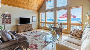 A spacious living room with a large TV, wooden ceiling, and large windows offering a view of mountains. - Breathtaking Mountain & Lake Views! (Dayton)