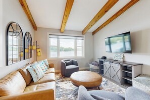 A modern living room with a leather sofa, a grey armchair, and a round ottoman. There is a large window with a view of greenery outside, a flat-screen TV mounted on the wall, and a wooden ceiling with exposed beams.