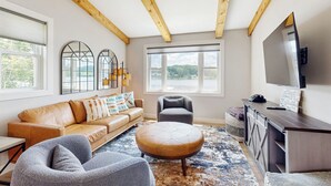 A living room with a sofa, ottoman, armchair, and a TV on a wooden cabinet. Large windows offer a view of the outdoors. - 3 BR Lakefront House w/ Private Dock (Lodi)