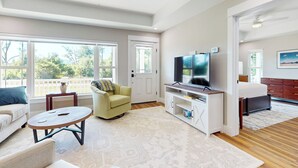 Ein geräumiges Wohnzimmer mit großem Fernseher, einer Couch, einem Couchtisch und einem Balkon mit Blick auf Bäume.