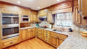 A modern kitchen with wooden cabinets, stainless steel appliances, granite countertops, and a window with curtains. - 4BR Waterview Home w/ Pool, Fire Pit & Fishing (Griffin)