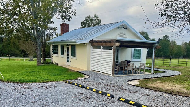 A small, single-story house with a garage, a covered porch with chairs, and a gravel driveway. - Windmill Trails Farm - Barn Loft and Dairy Cottage (Tylertown)