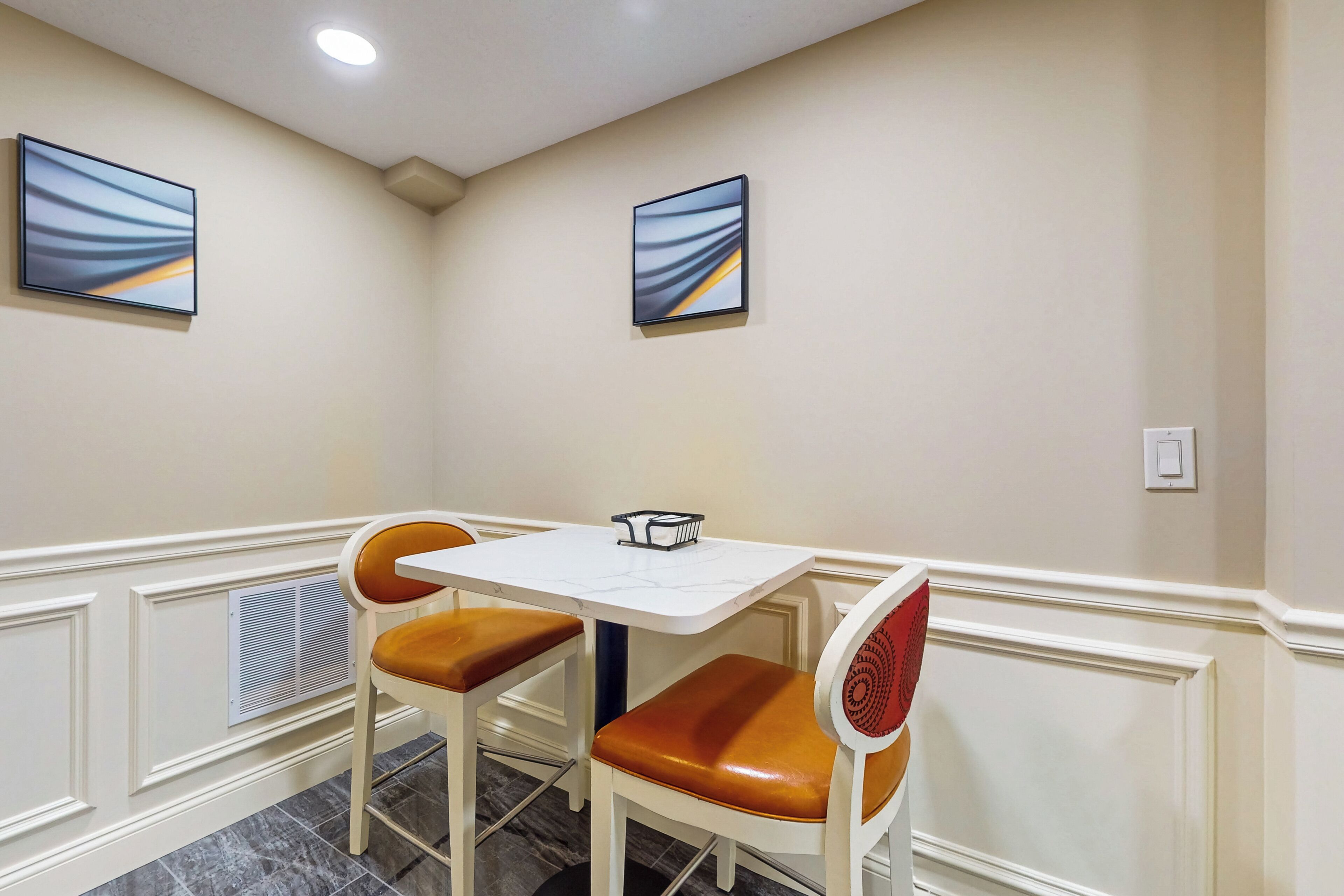 A dining area with a white table and two chairs with leather seats, a white wall with two framed abstract paintings, and a ceiling light.