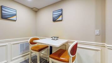 A dining area with a white table and two chairs with leather seats, a white wall with two framed abstract paintings, and a ceiling light.