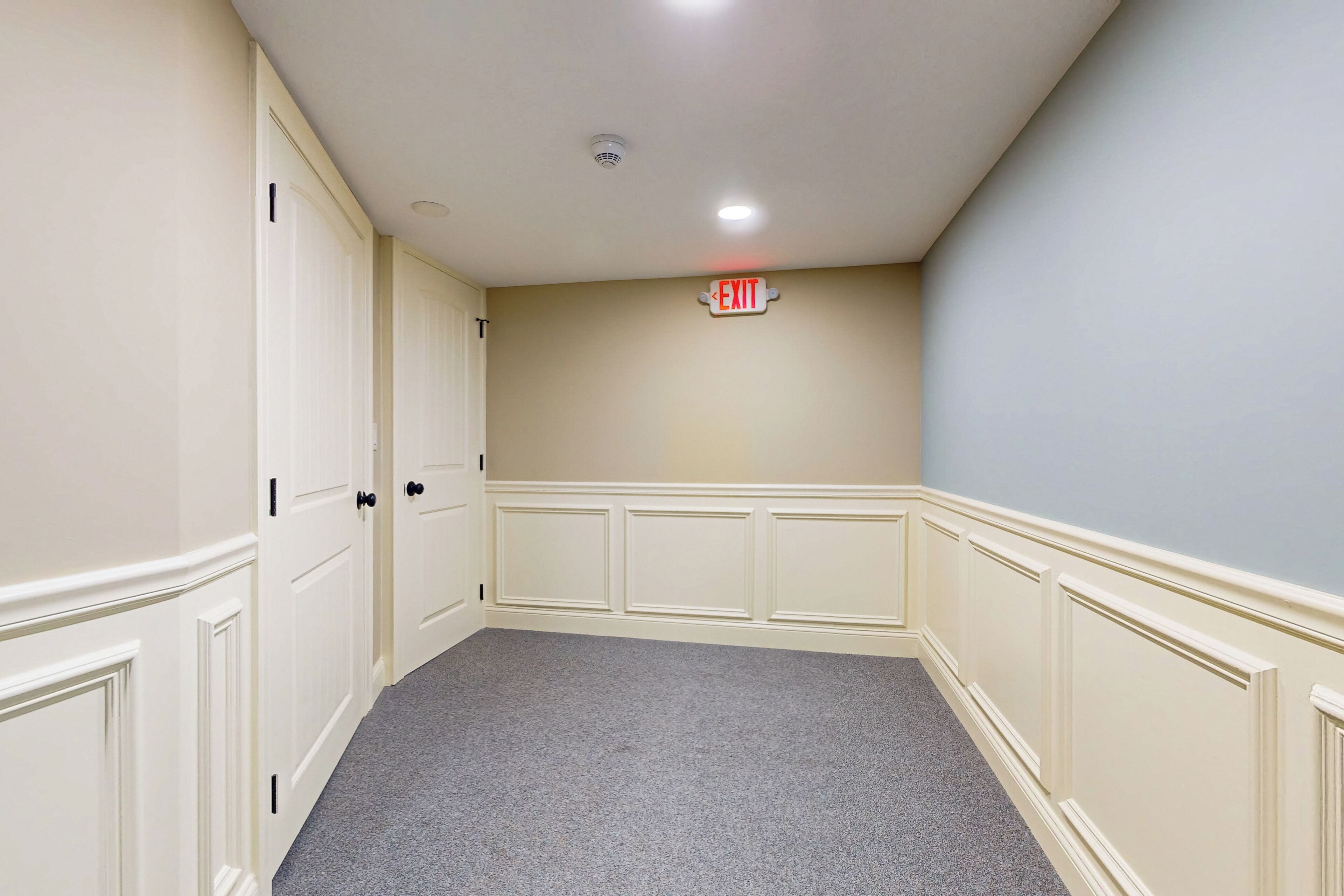 A hallway with cream-colored walls, a grey carpet, and white doors. An exit sign is visible on the wall.