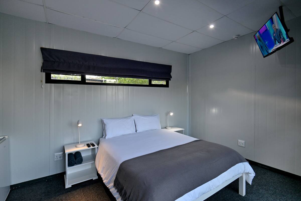 A bedroom with a large bed, a nightstand with a lamp, a TV mounted on the wall, and a window with blinds.