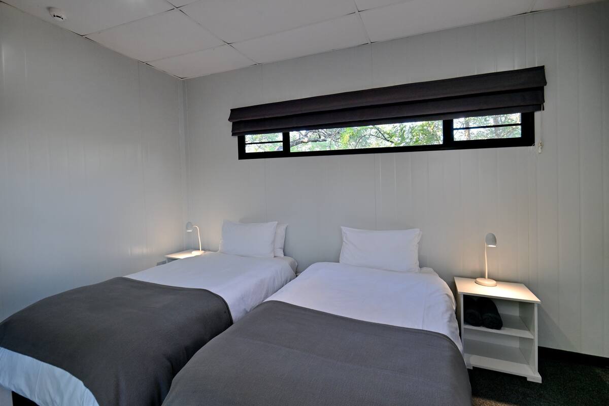 A room with two beds, a window with blinds, a nightstand with a lamp, and a shelf with shoes.