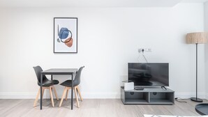 Un salon moderne comprenant une table à manger, deux chaises, un téléviseur à écran plat, une lampe et une œuvre d’art encadrée accrochée au mur.