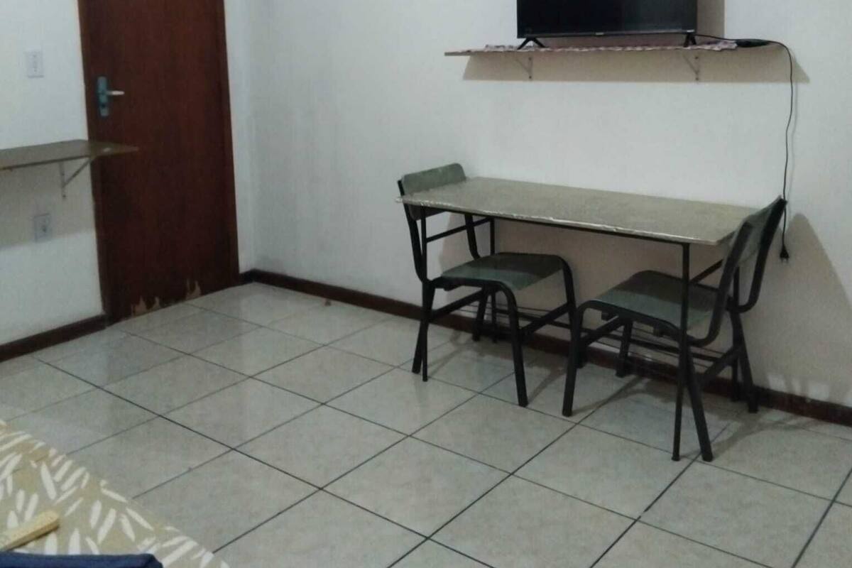 Una habitación pequeña con una puerta de madera, un televisor en un estante, una mesa con sillas y suelo de baldosas.