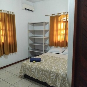 A small, clean room with a bed, a white shelf unit, and a window with curtains. - Pousada mambucaba  (Angra dos Reis)