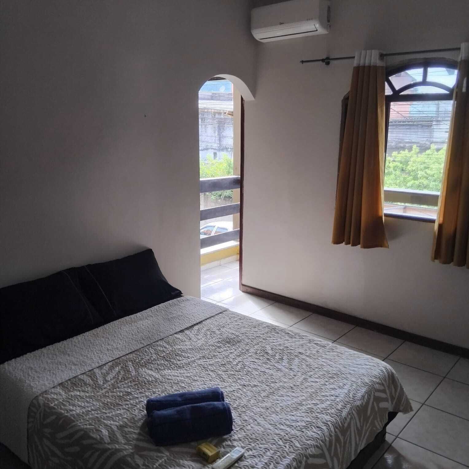 A bedroom with a bed, a window with curtains, an air conditioning unit, and a view of greenery outside.