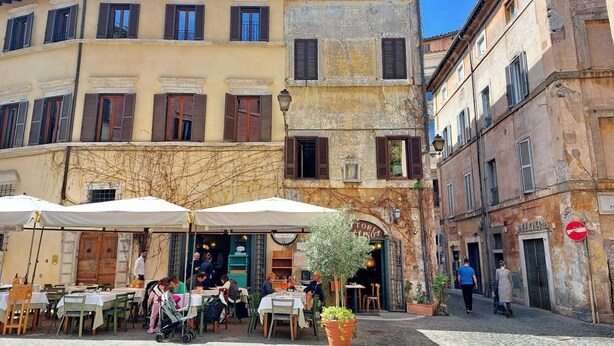 An outdoor café with tables and chairs, surrounded by historic buildings with shuttered windows and a cobblestone street. - Luxury home near Piazza Venezia (Roma)