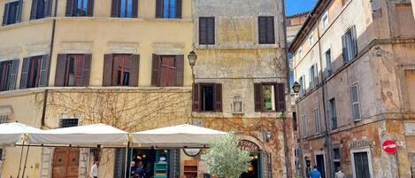 An outdoor café with tables and chairs, surrounded by historic buildings with shuttered windows and a cobblestone street.