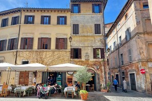 Um café ao ar livre com mesas e cadeiras, rodeado por edifícios históricos com janelas protegidas por persianas e uma rua de calçada.