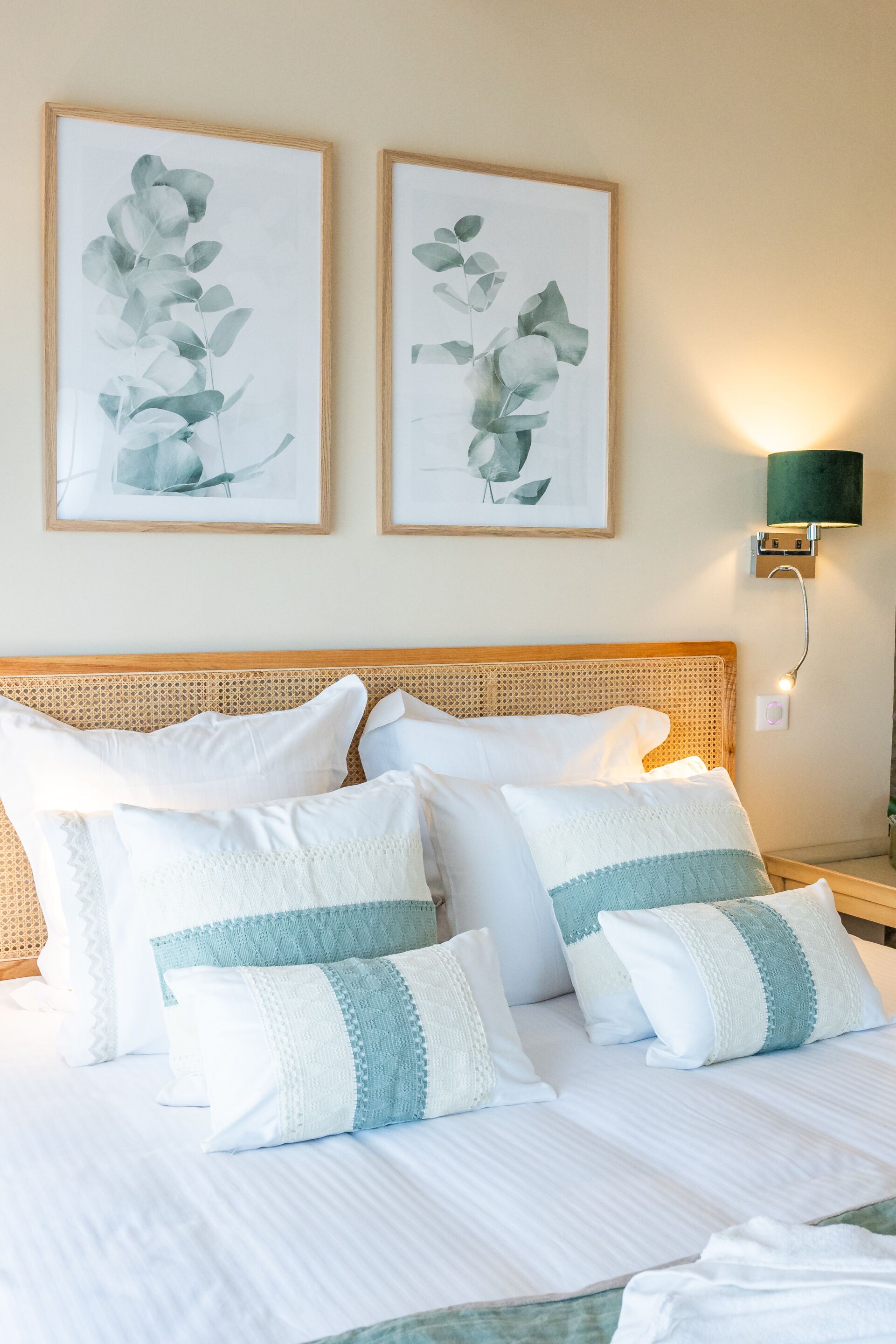 Een net opgemaakt bed met witte en lichtblauwe kussens, een geweven hoofdbord en twee ingelijste botanische prints aan de muur.