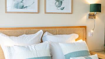 Een net opgemaakt bed met witte en lichtblauwe kussens, een geweven hoofdbord en twee ingelijste botanische prints aan de muur.