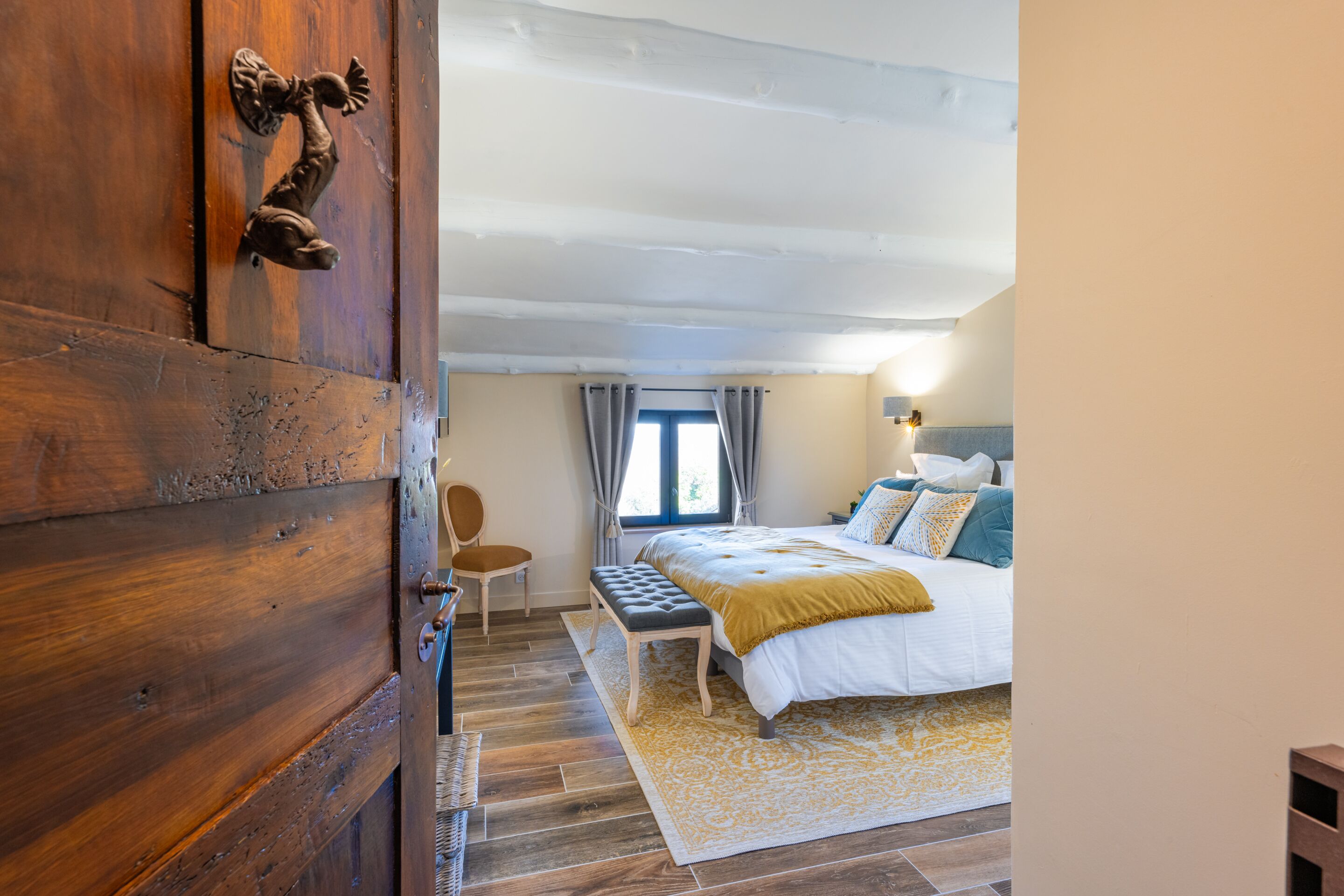 A bedroom with a wooden door, a bed with a white and yellow bedspread, a bench, and a window with curtains.