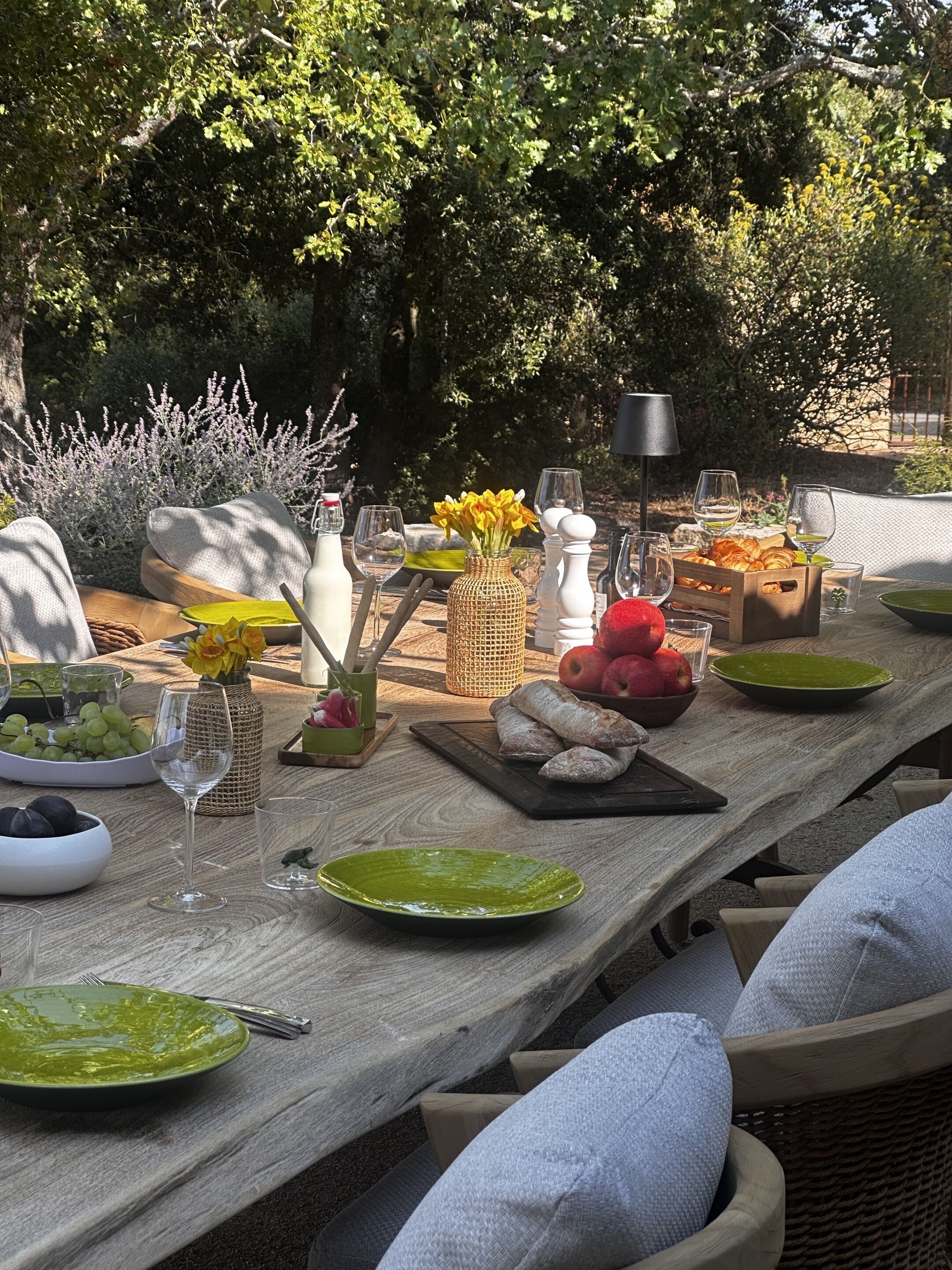 Mesa de comedor al aire libre con platos, vasos y comida, rodeada de follaje y una lámpara.