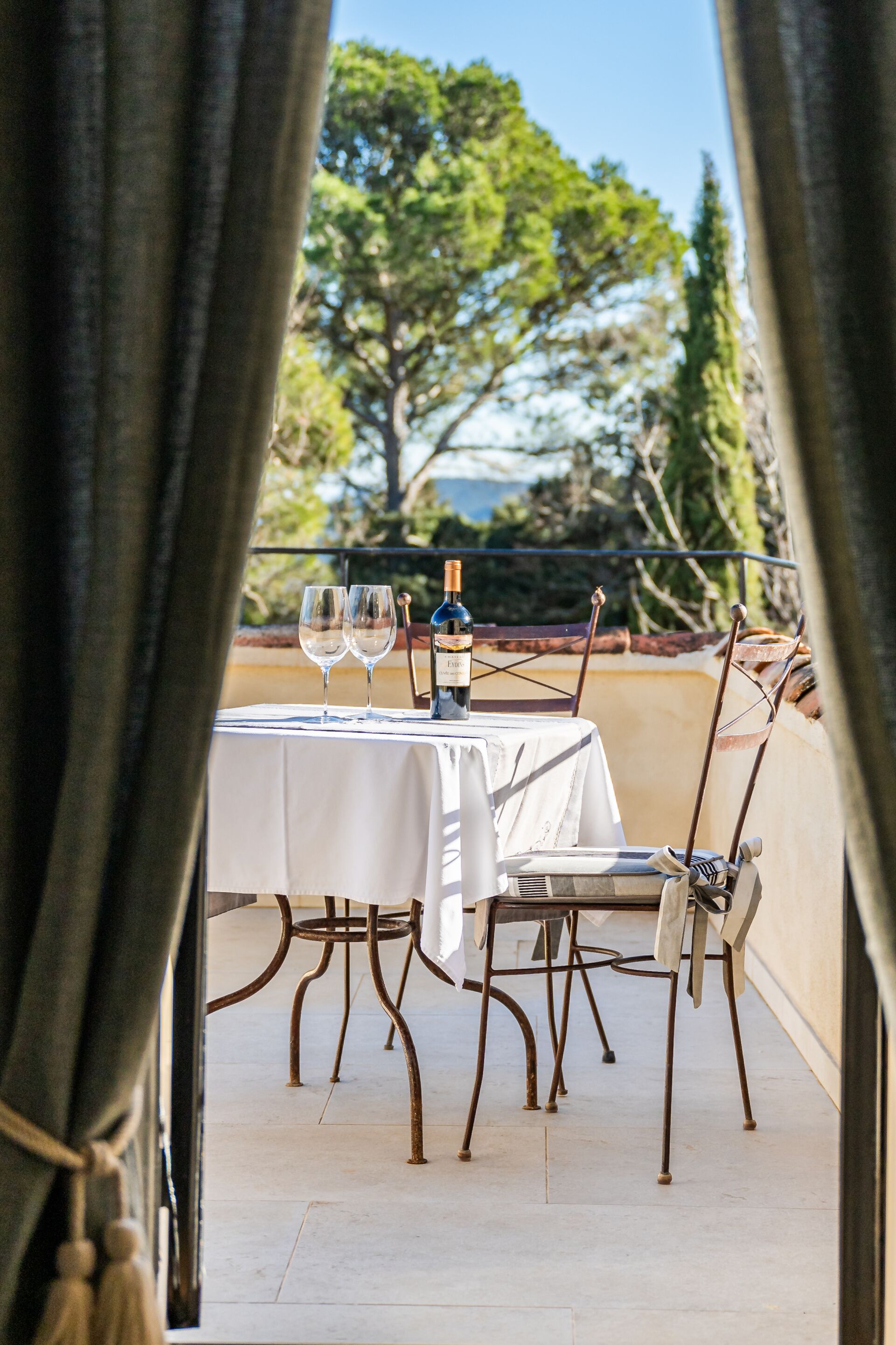 A table set for two with a bottle of wine and two glasses, overlooking a garden with trees.