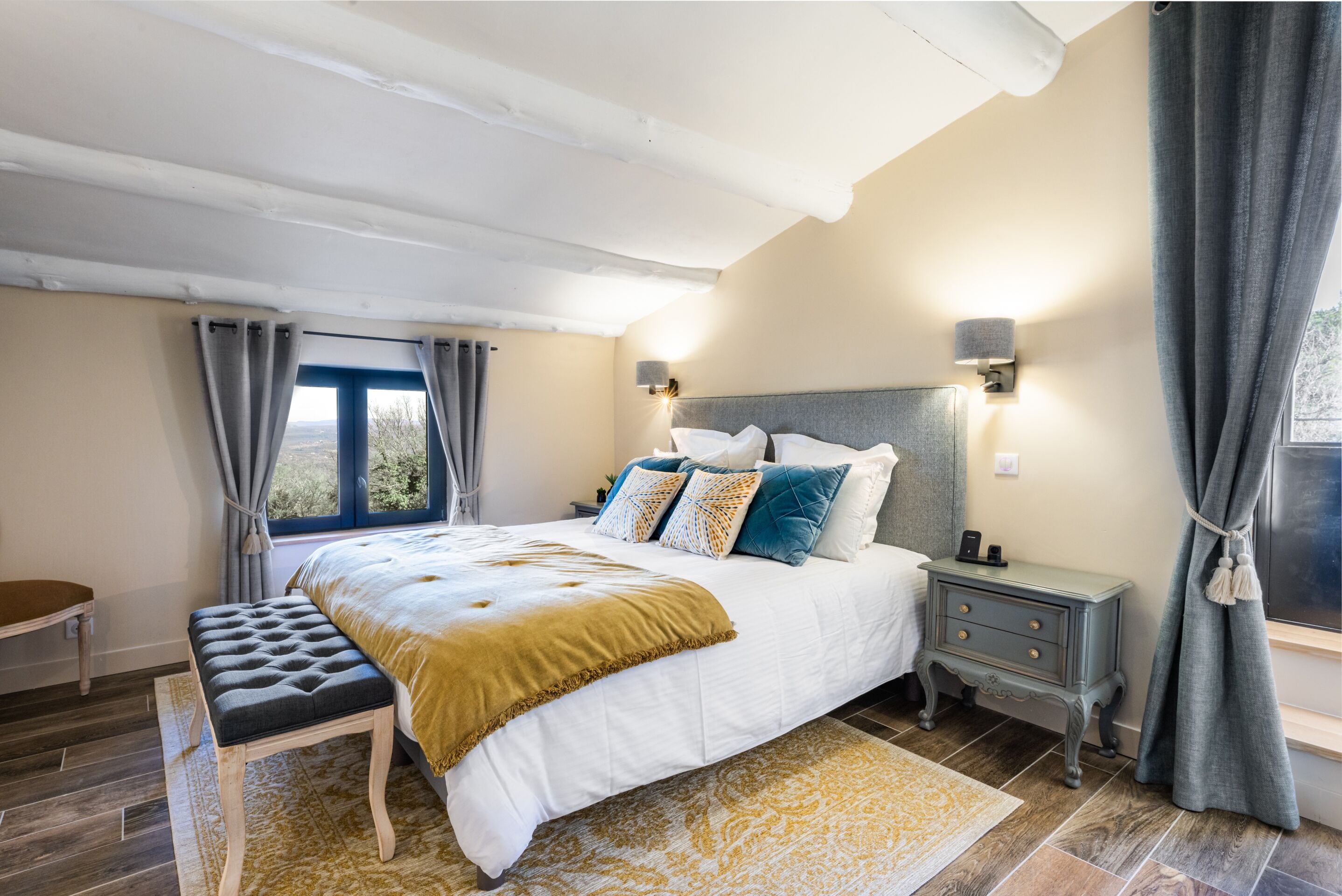 A bedroom with a large bed, bedside tables, a bench, and a view of the outdoors.