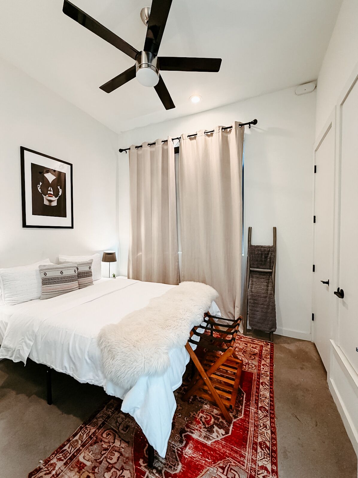 Un dormitorio con cama, ventilador de techo, cuadro enmarcado, ventana con cortinas, silla de madera y alfombra estampada.