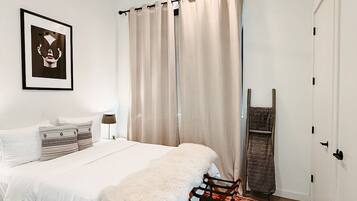 Un dormitorio con cama, ventilador de techo, cuadro enmarcado, ventana con cortinas, silla de madera y alfombra estampada.