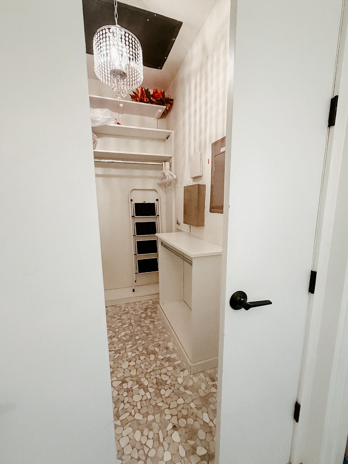 A narrow hallway with a tiled floor, a white door, a small window, and a chandelier.