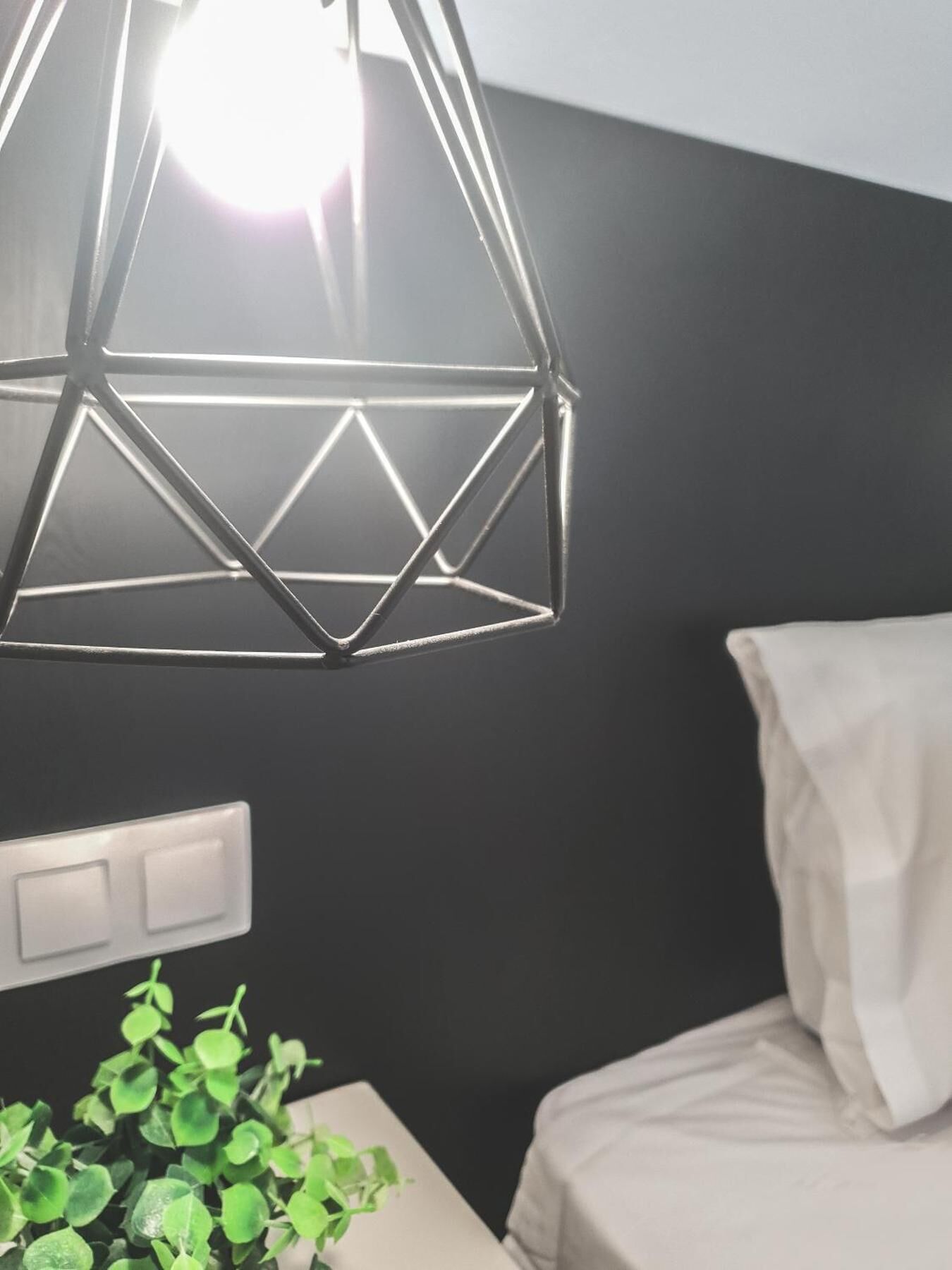 Een kamer met een geometrische hanglamp, een nachtkastje met een groene plant, en een wit bed.