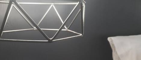Une chambre avec un luminaire suspendu aux formes géométriques, une table de chevet ornée d’une plante verte et un lit blanc.