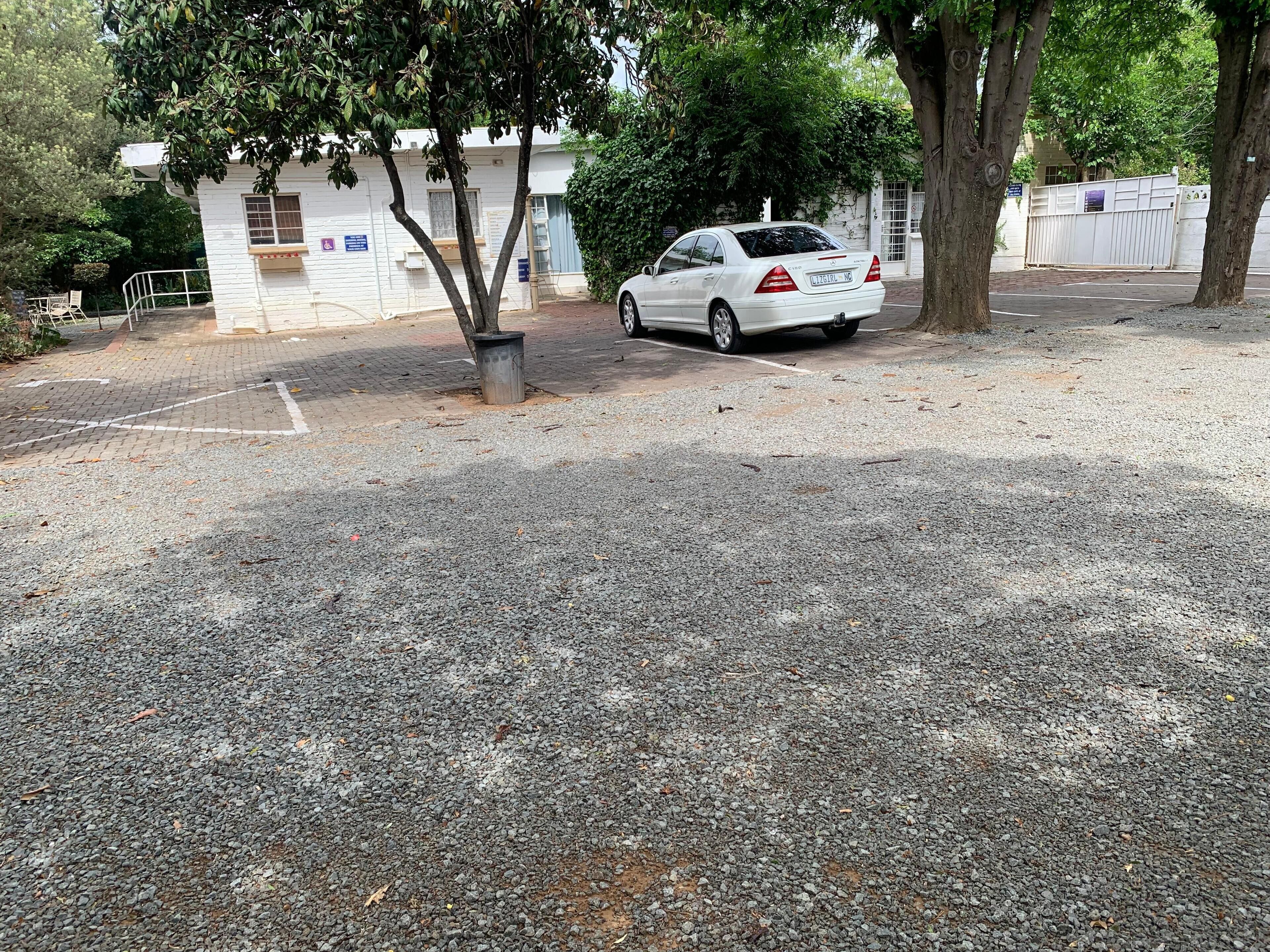 Un coche blanco estacionado en un área de grava, con árboles y un edificio al fondo.