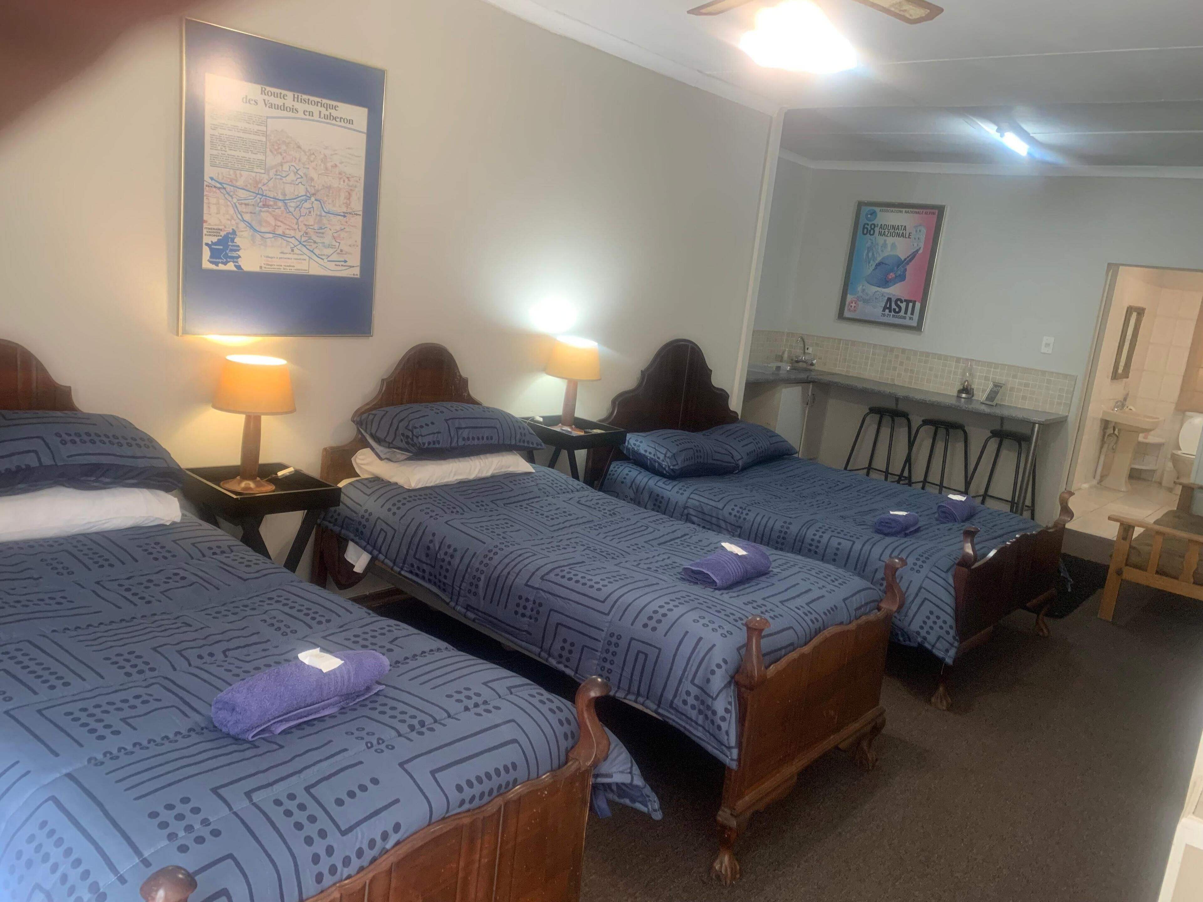 A hotel room with three beds, each with a patterned bedspread and pillows, a small table with a lamp, a map on the wall, and a bathroom with a sink and mirror.