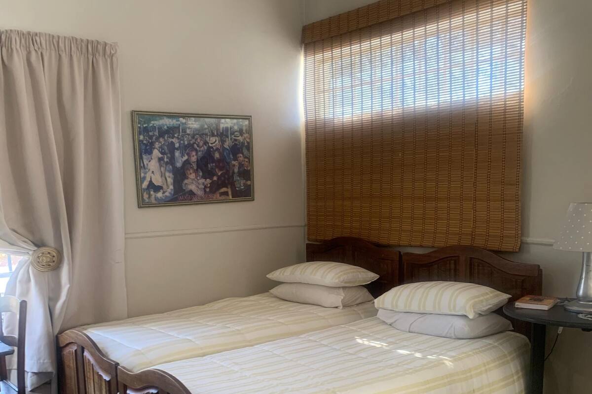 Un dormitorio con cama, cabecera de madera, mesita de noche con lámpara, una silla y ventana con persianas.