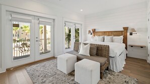 A bedroom with a bed, a sofa, two ottomans, a ceiling fan, and a view of palm trees through the sliding glass doors. - Sandy Cheeks - Stunning 4 Bedroom with 4 bikes & seconds to the Resort Pool! (Panama City Beach)