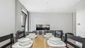 A modern dining room with a long table set for six, grey upholstered sofas, and a large flat-screen TV mounted on the wall. - Modern & Spacious Studio Apartment in Leicester (Leicester)
