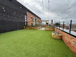 Eine Dachterrasse mit künstlichem Rasen, Holzpflanzgefäßen und Lichterketten.