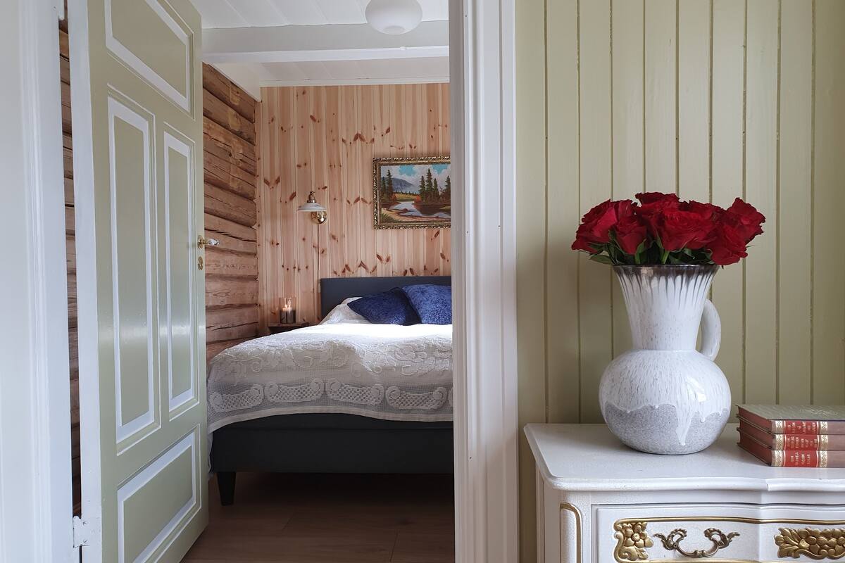 Un dormitorio con una cama, una mesita de noche, un florero con flores rojas y un aparador con libros.
