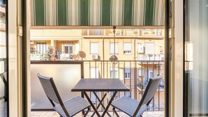 Een balkon met een tafel en twee stoelen, een gestreept zonnescherm en uitzicht op een woongebouw.